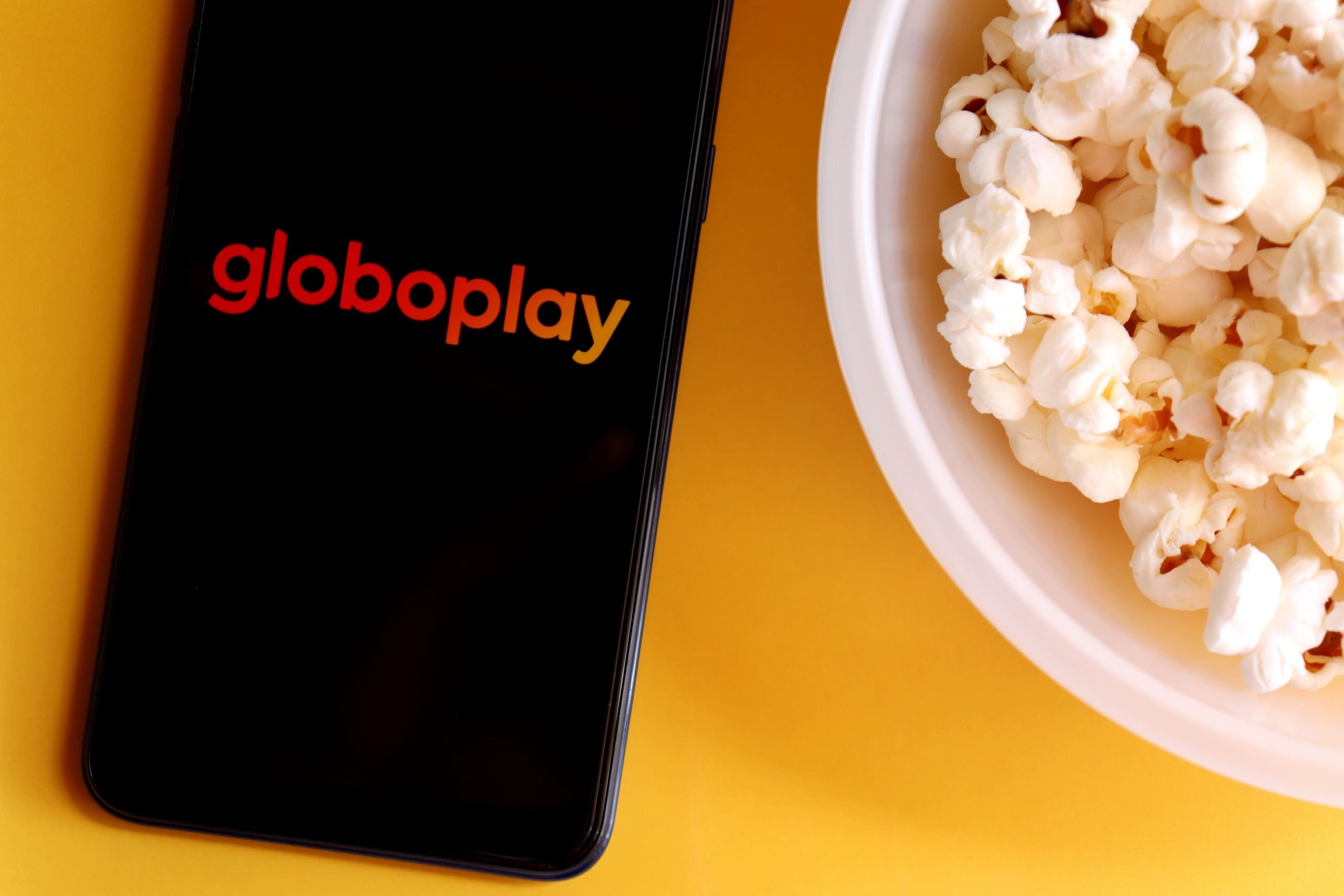 Fundo amarelo com um telefone e pipoca pronta para começar a ver os melhores filmes do Globoplay