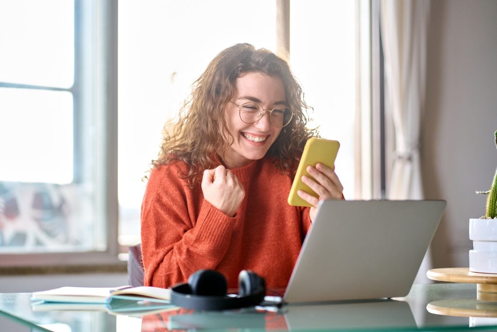 mulher feliz com celular na mão e notebook aberto