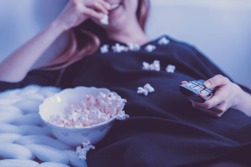 Mulher deitada assistindo TV e comendo pipoca.