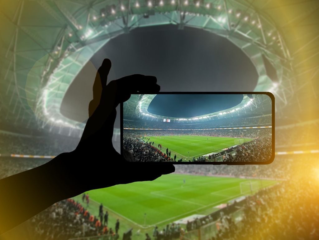 mão segurando celular com estádio de futebol ao fundo