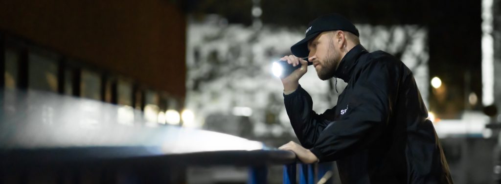 Security Guard Walking Building Perimeter With Flashlight At Night