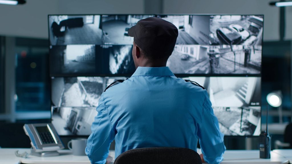 Back view of African-American security operator use computer with screens showing surveillance camera footage.